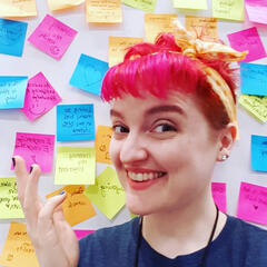 A headshot of Margaret Mae. She's a smiling white woman with bright pink hair stending in front of a wall of brightly colored postit notes